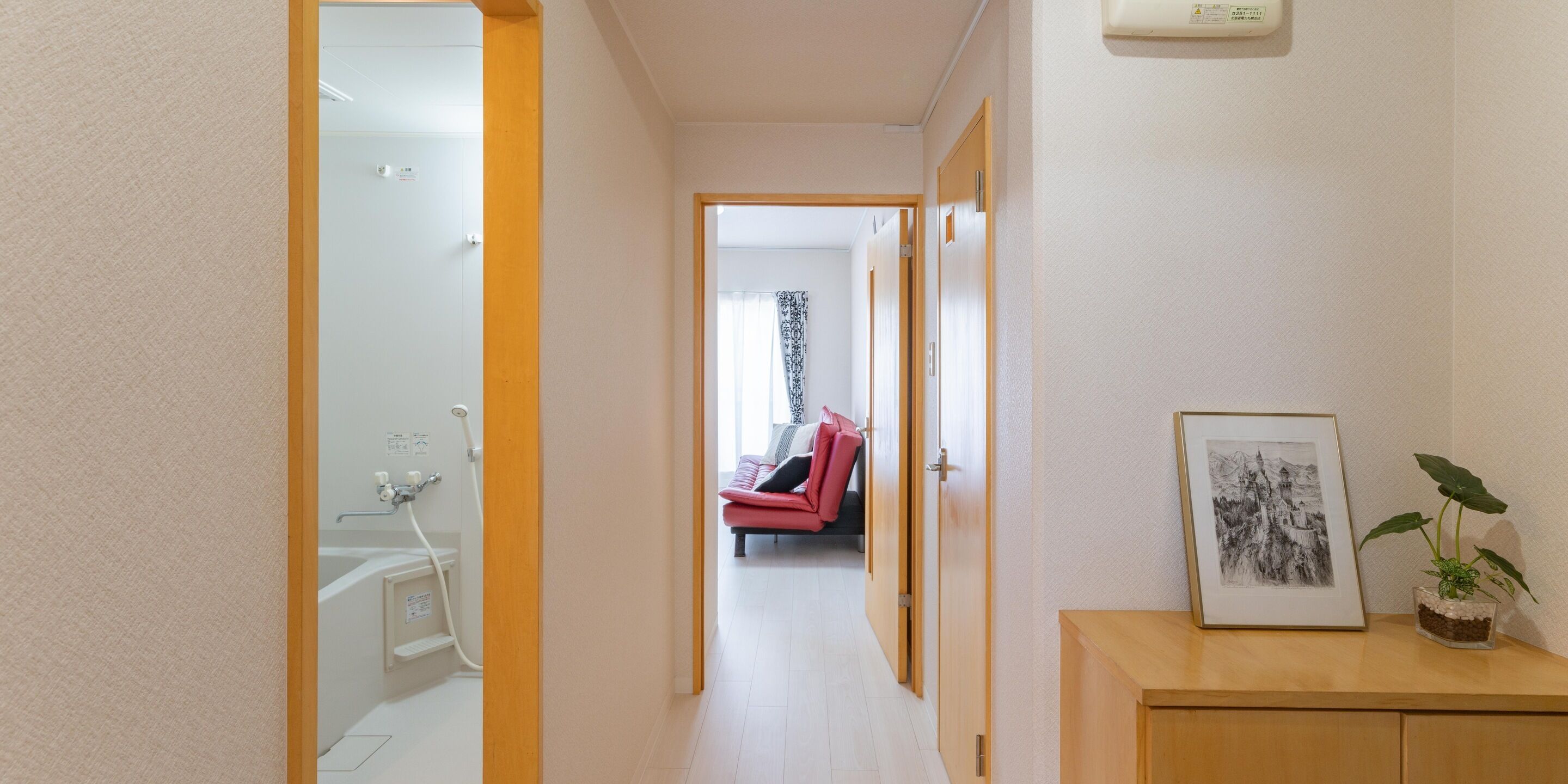 Un couloir mène à une salle de bain équipée d’une douche, où l’on trouve une chaise rouge et un tableau encadré posé sur un meuble en bois.