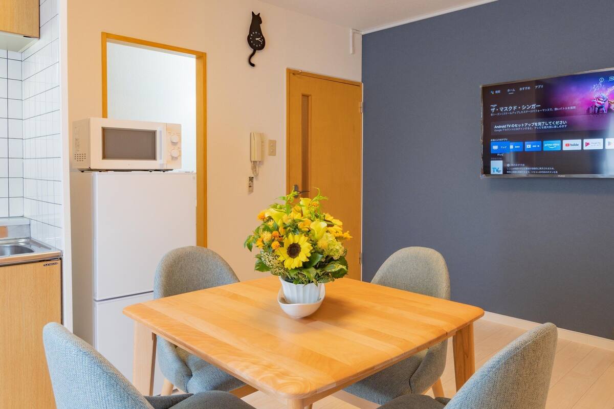 Uma cozinha moderna com armários brancos, um micro-ondas e uma mesa de jantar de madeira com duas cadeiras. Um vaso de girassóis está sobre a mesa.