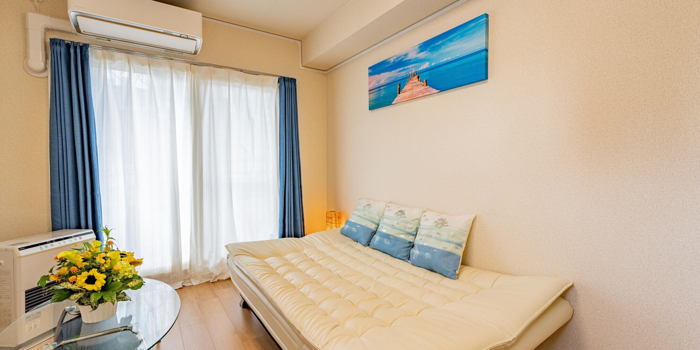 A hotel room with a bed, air conditioning unit, curtains, a painting, and a flower vase.