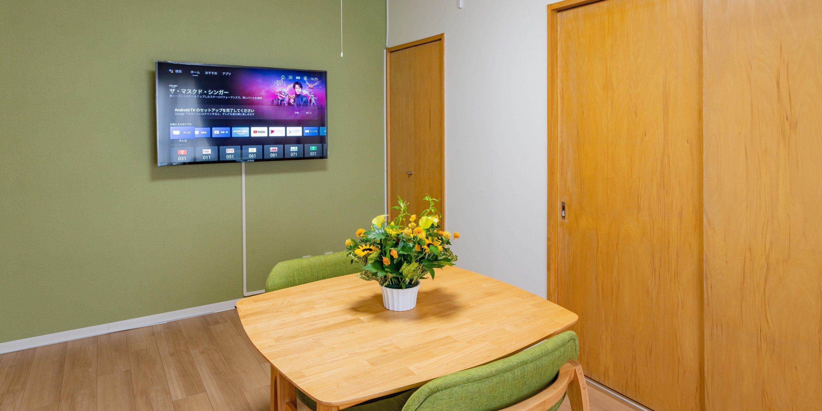 Een vergaderruimte met een houten tafel, groene stoelen, een potplant en een flatscreen-tv aan de muur waarop een video wordt afgespeeld.