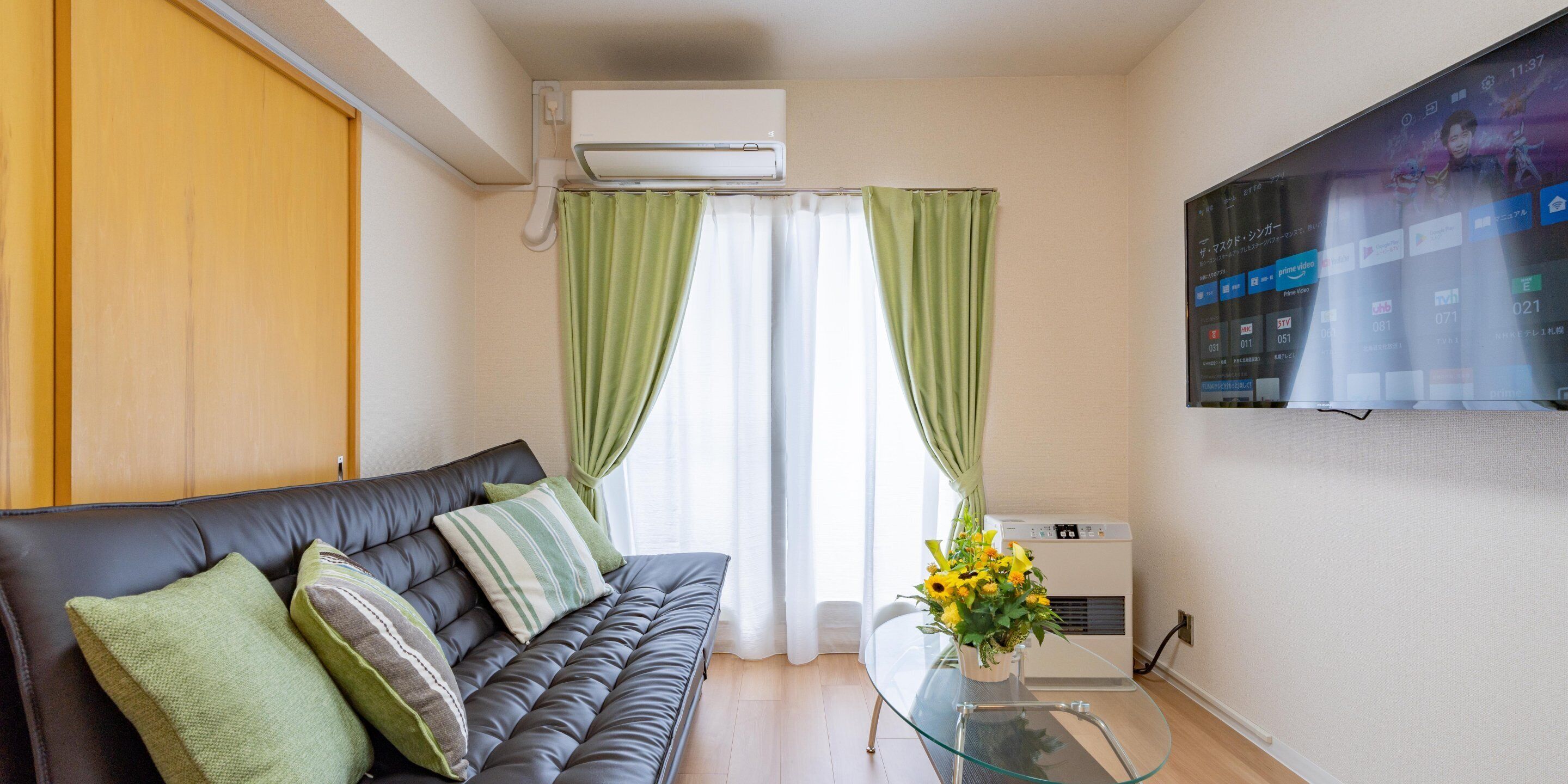A modern living room with a leather sofa, green curtains, a glass coffee table, and a mounted TV.