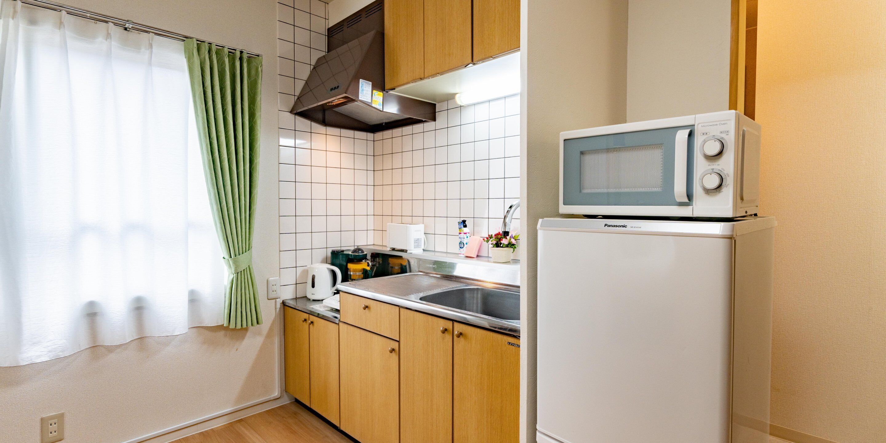 A compact kitchen with wooden cabinets, a white refrigerator, a microwave, a sink, and a window with a green curtain.