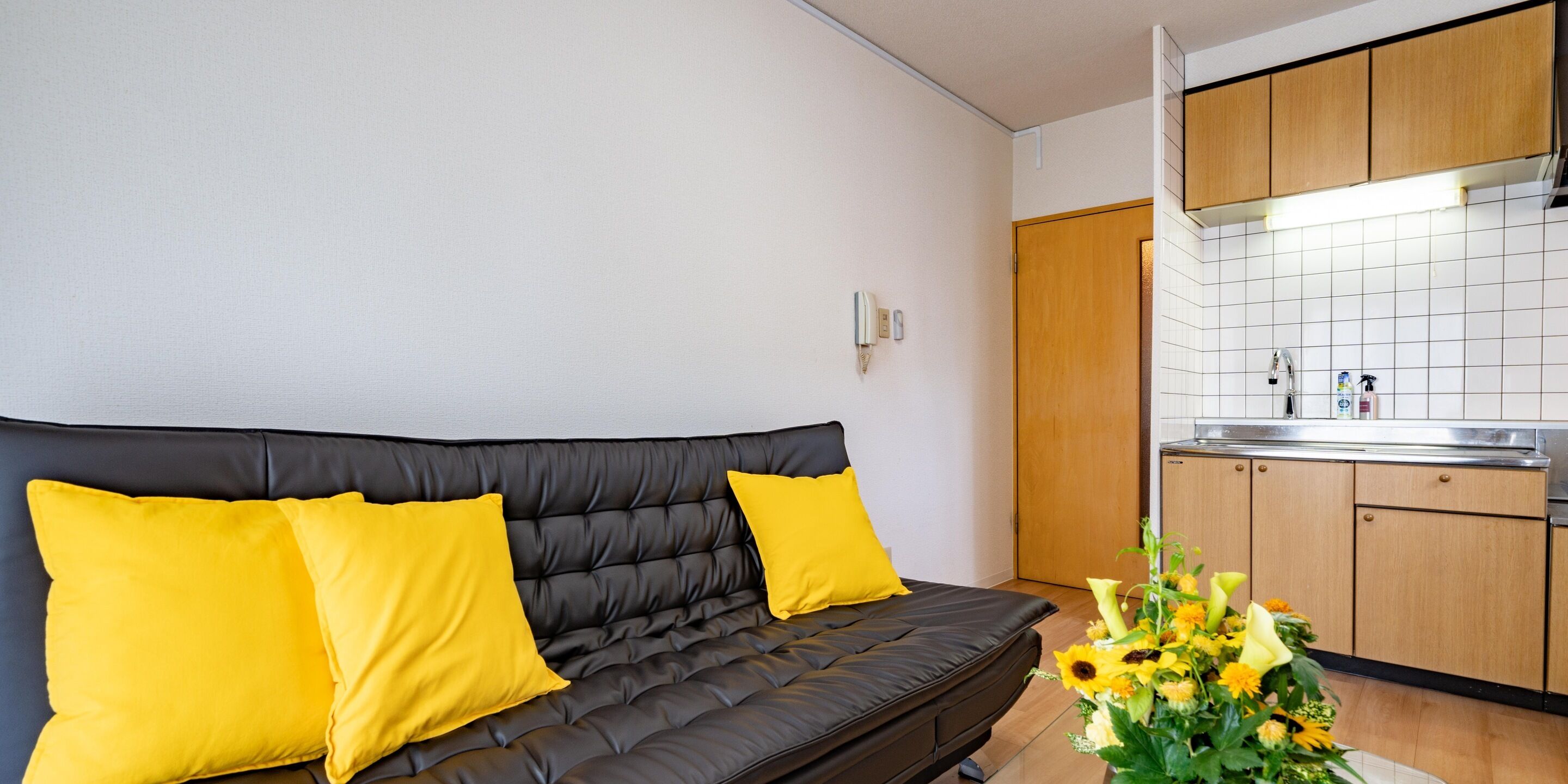 Un espace de vie moderne avec un canapé en cuir noir, des coussins jaunes, un coin cuisine doté de meubles en bois et un mur recouvert de carreaux.