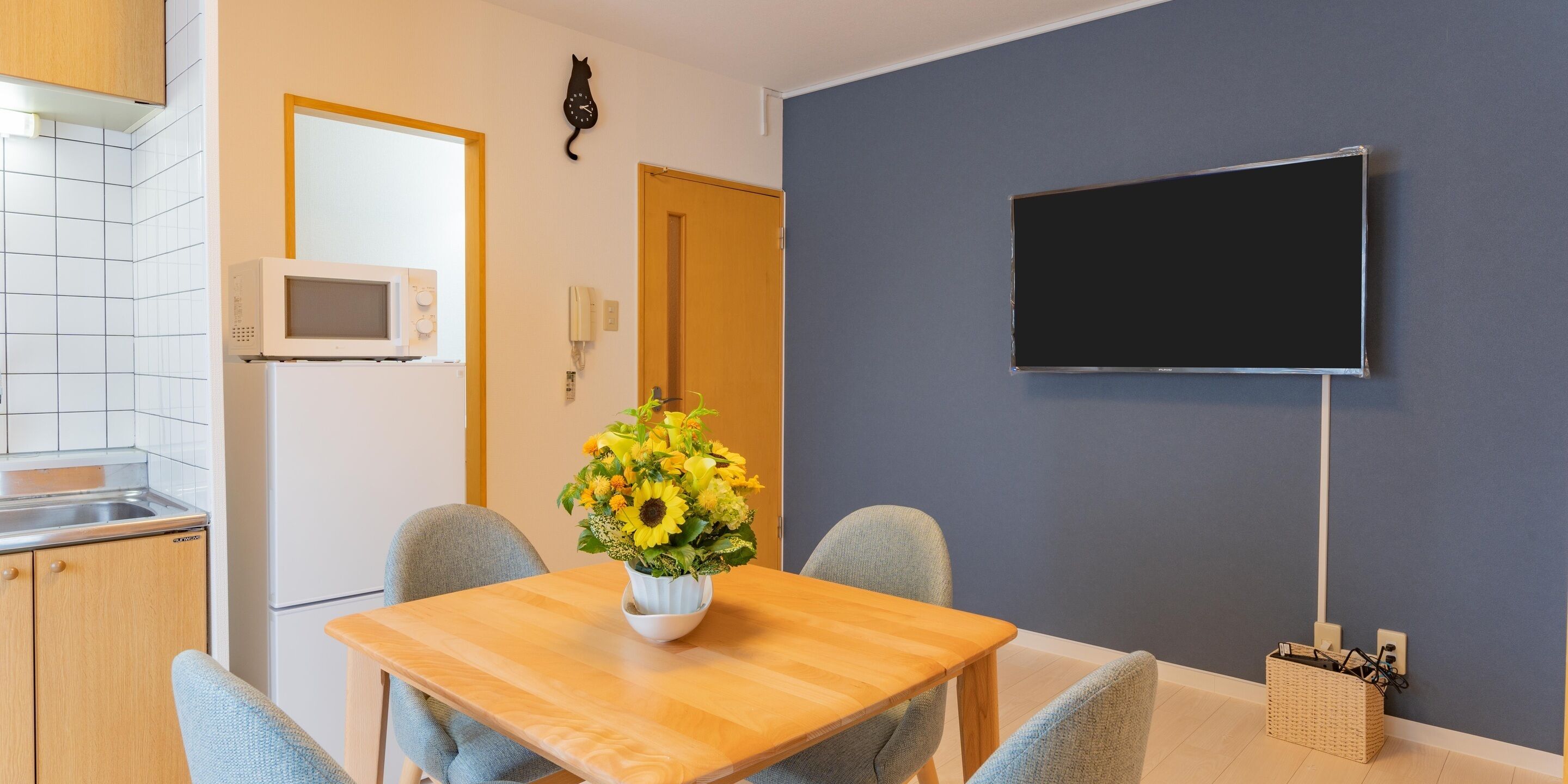 Ein Essbereich mit einem Holztisch, zwei Stühlen, einer Vase mit Sonnenblumen, einer Mikrowelle, einem Telefon und einem an der Wand befestigten Fernseher.