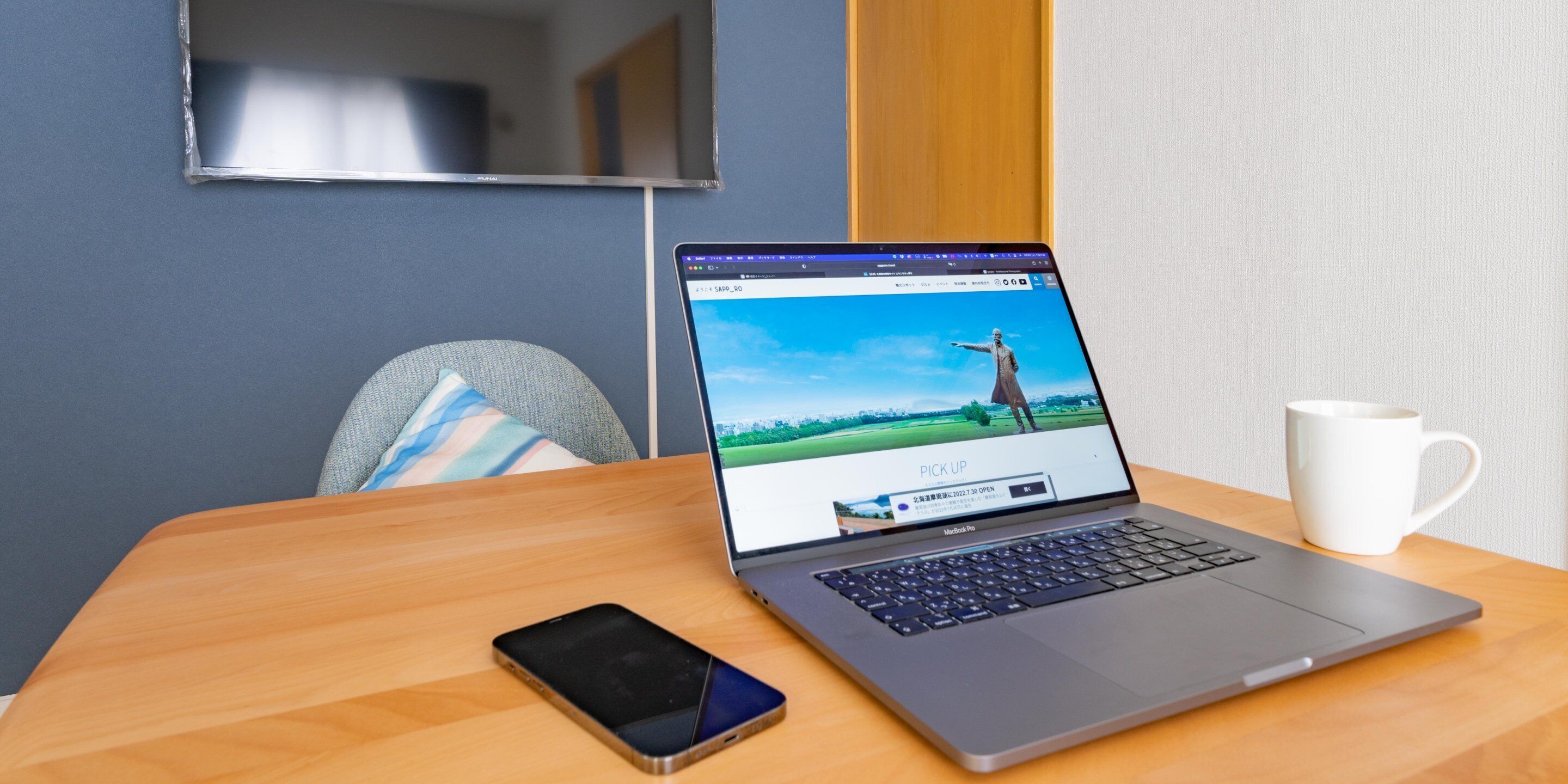 A laptop on a wooden table with a smartphone beside it, a white mug, and a chair with a striped cushion.