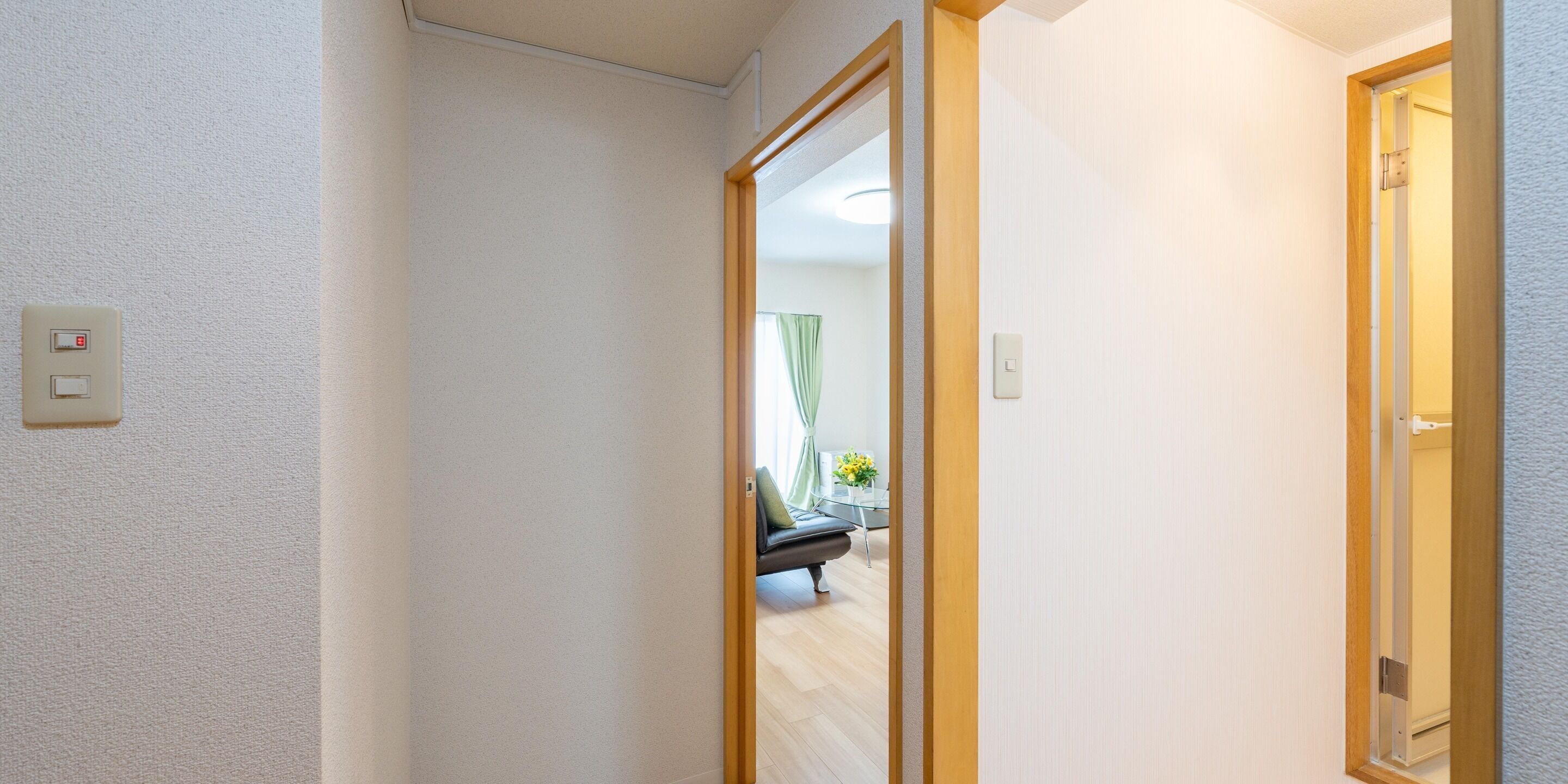 Un pasillo con una puerta que da a una habitación, un interruptor de luz y parte visible de una cama con un almohadón verde.