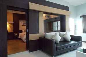 A modern hotel room with a dark-colored sofa, a wooden accent wall, and a large window providing natural light. - Eastpana Hotel (Si Racha)