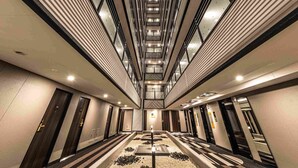 Un pasillo moderno de hotel con una escalera central, varios ascensores y un techo alto con tragaluces.