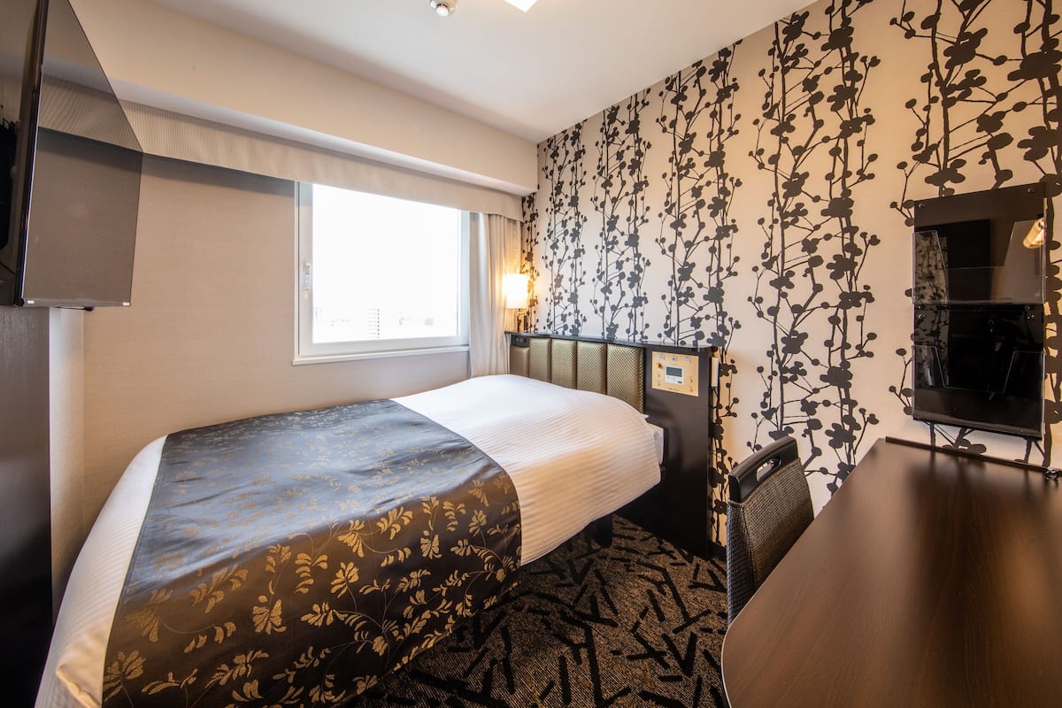 A hotel room with a bed, a desk, a chair, a TV, a window, and a wall with a floral pattern.