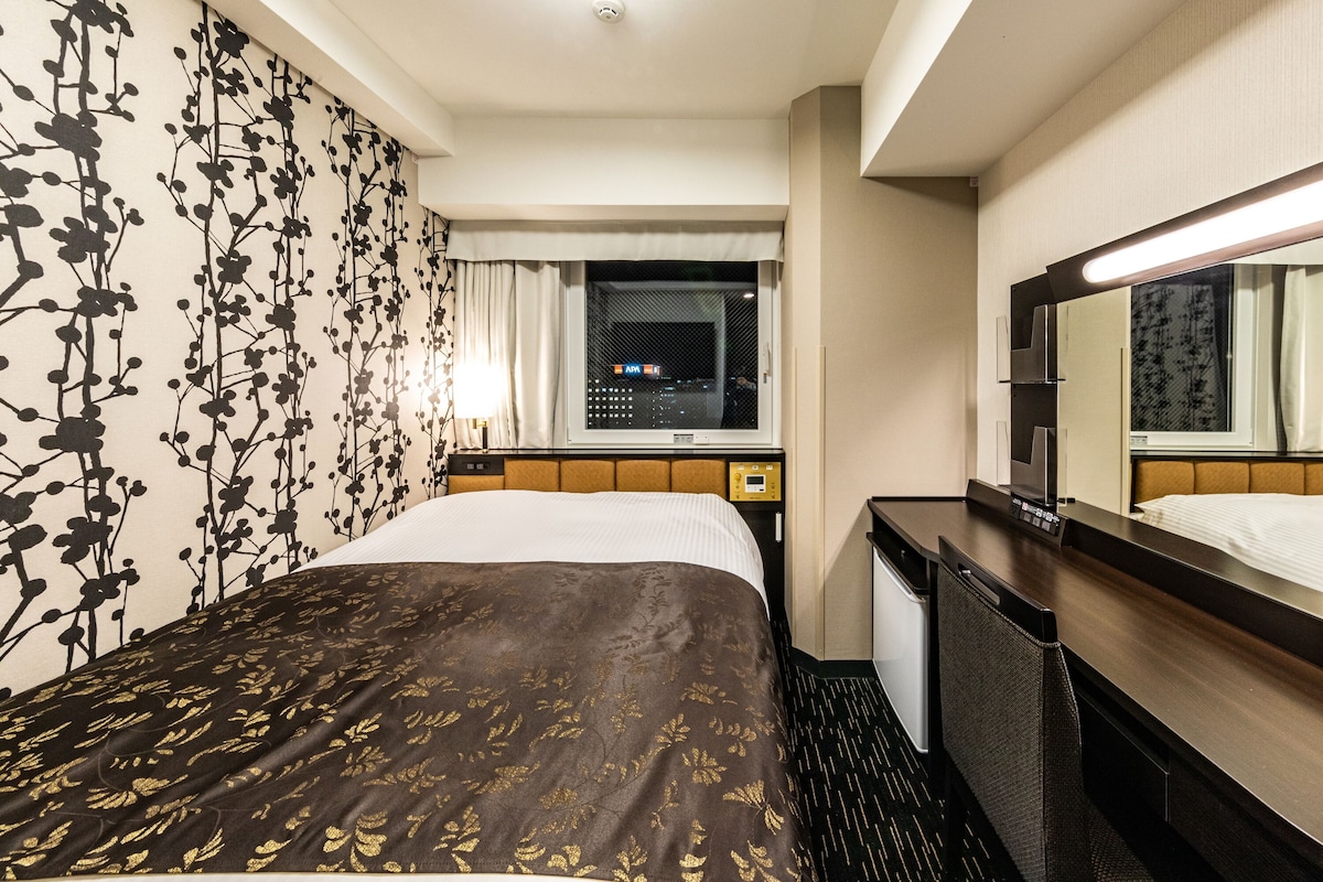 Ein Hotelzimmer mit Bett, Schreibtisch, Spiegel, Fenster mit Blick auf die Stadt und Wand mit Blumenmuster.