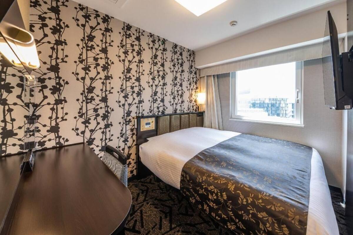 A hotel room with a bed, a desk, a chair, a window with a city view, and a flat-screen TV.
