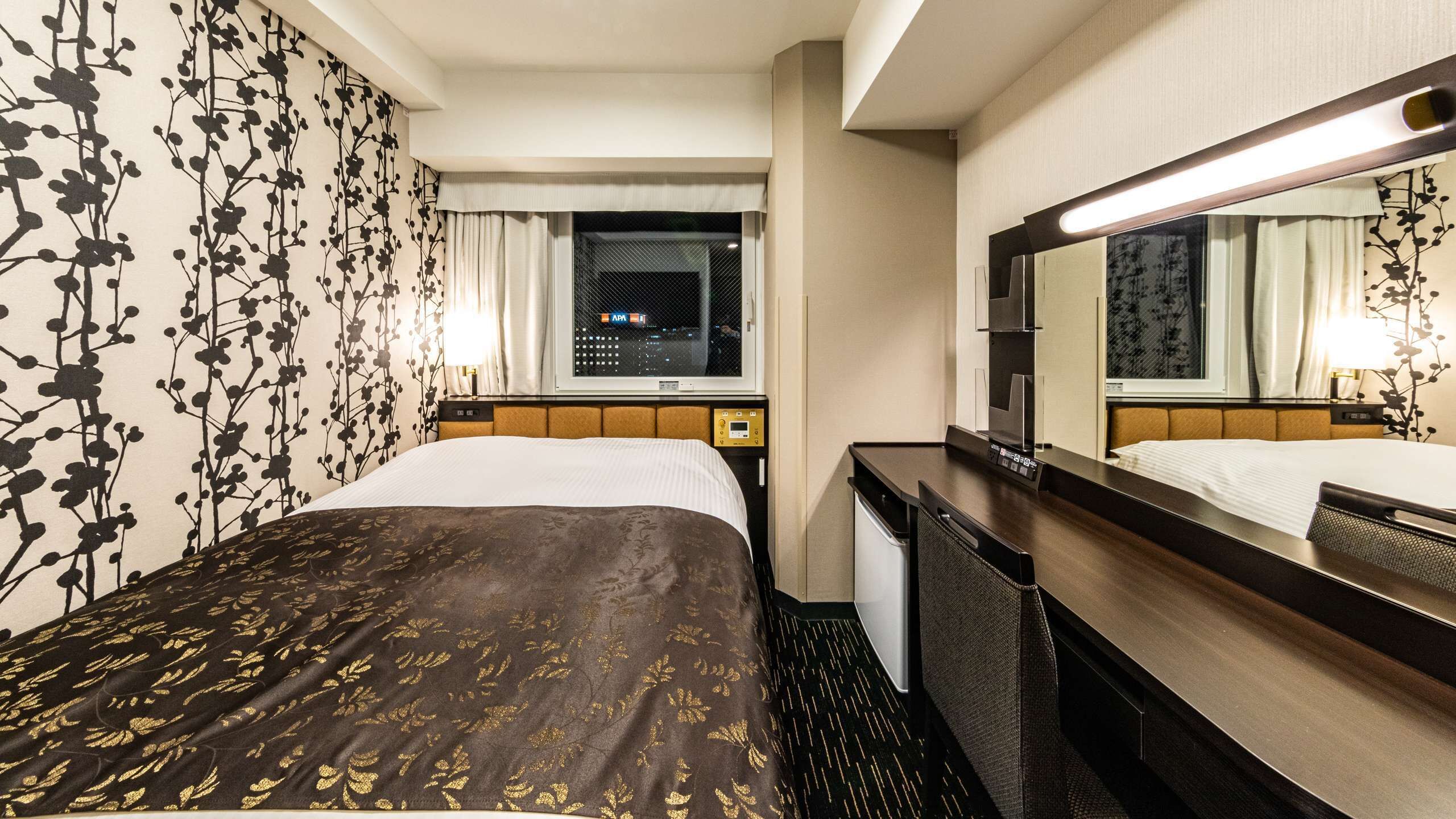 A hotel room with a bed, a nightstand, a mirror, and a window with a view of the city at night.