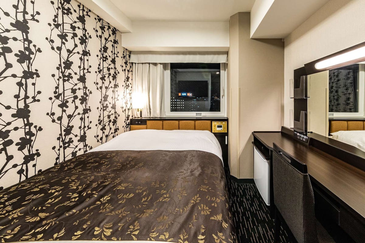 A hotel room with a bed, a nightstand, a mirror, and a window with a view of the city at night.