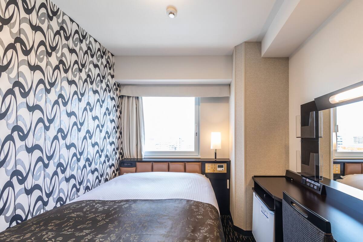 A hotel room with a bed, a desk, a chair, a TV, a window, and a wall with a patterned design.