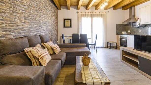 A cozy living room with a stone wall, a brown sofa, a wooden coffee table, and a kitchen area with a stove and cabinets.