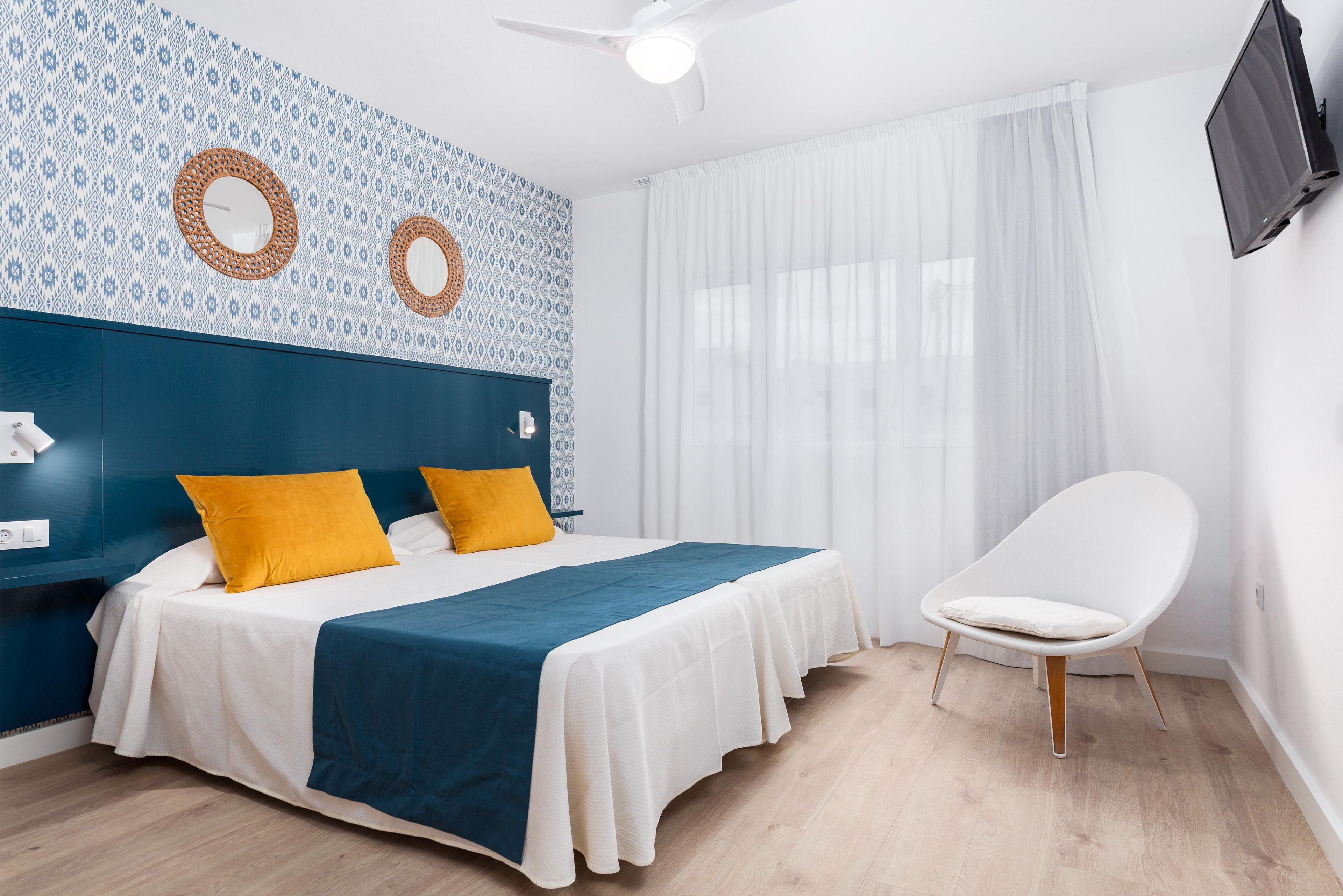 Un dormitorio con cama, una silla, ventilador de techo y televisor montado en la pared.