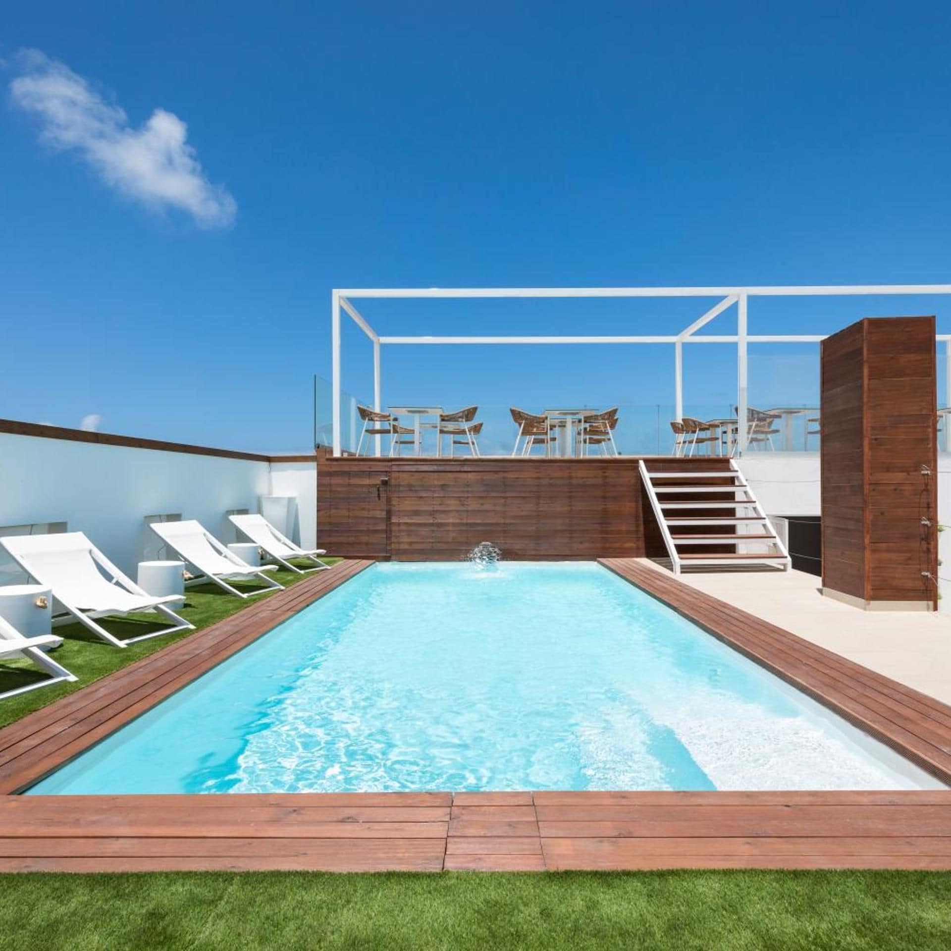 A rooftop pool with a wooden deck, white lounge chairs, and a glass railing.