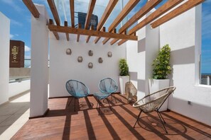 A covered outdoor area with wooden beams, two blue chairs, a wicker chair, and decorative wall art. - Soul Surfer (La Oliva)