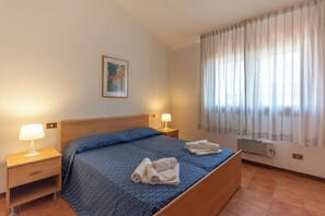 Hotellihuone, jossa on sänky, kaksi yöpöytää valaisimineen, seinään asennettu ilmastointilaite ja ikkuna, jossa on verhot.
