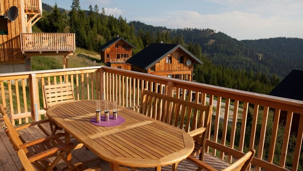 Terraza de madera con una mesa redonda y sillas, con vistas a una cadena montañosa boscosa.