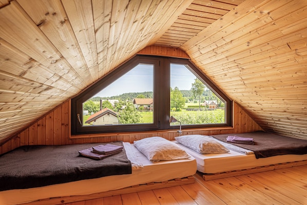 A cozy attic bedroom with two beds, wooden walls, and a large window offering a view of the countryside.