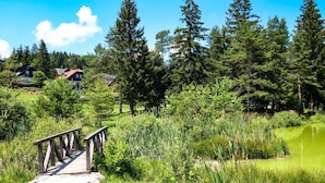 A wooden bridge leading to a pond surrounded by dense greenery and houses in the background. - Kraševec Lodge with Garden & Pool, Bloke, Slovenia (Bloke)