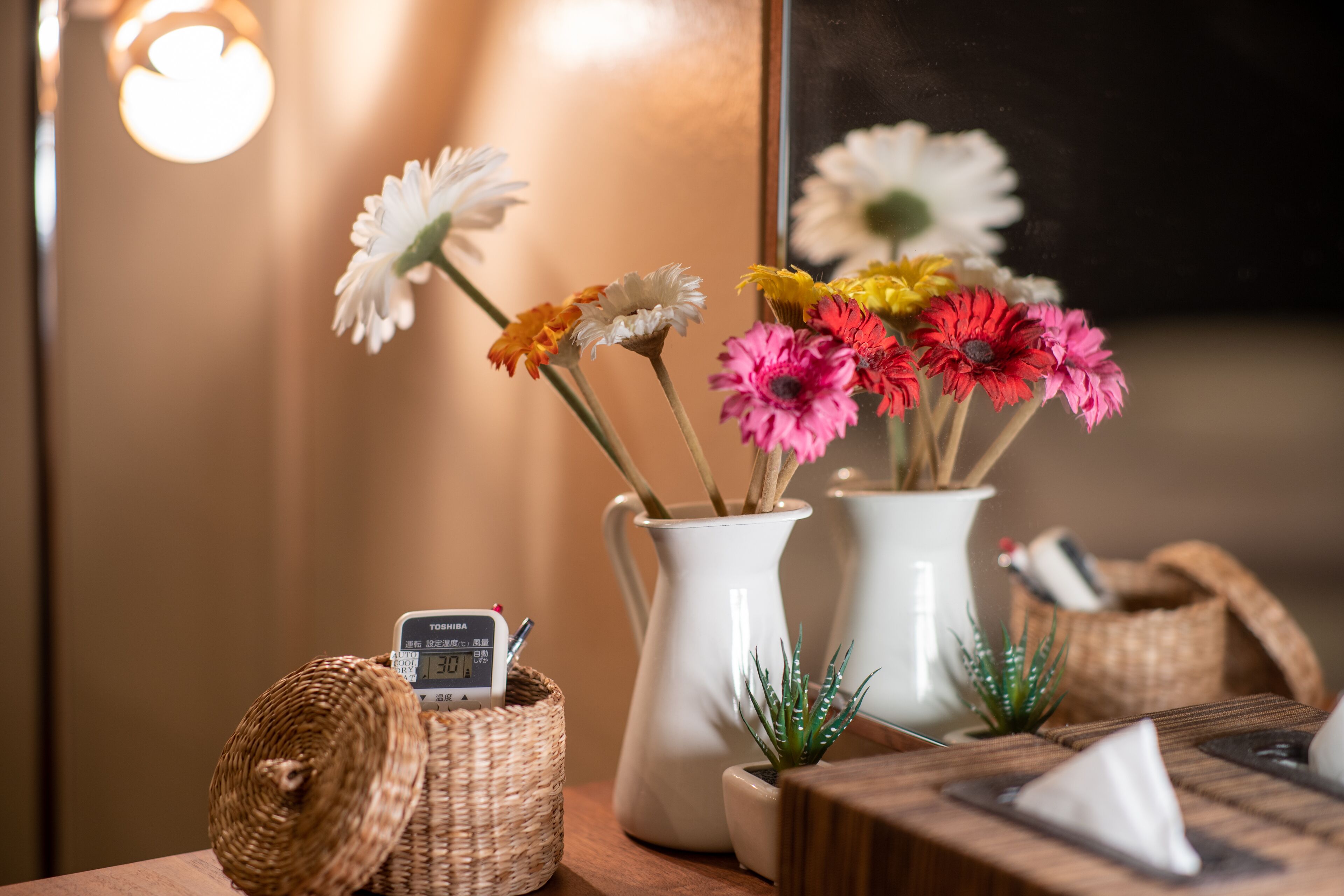 Une table en bois sur laquelle se trouve un vase blanc contenant des fleurs aux couleurs vives, une petite plante en pot et un panier tressé avec une horloge numérique.