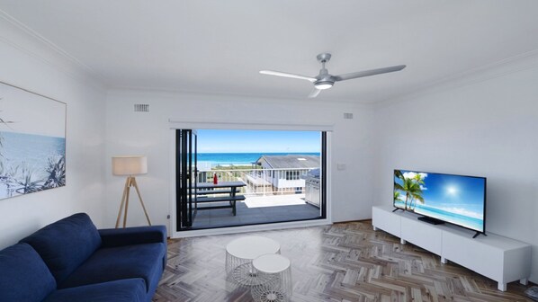 Un salon moderne avec un canapé bleu, un téléviseur posé sur un meuble blanc, un ventilateur de plafond et une vue sur l’océan par une porte-fenêtre coulissante.
