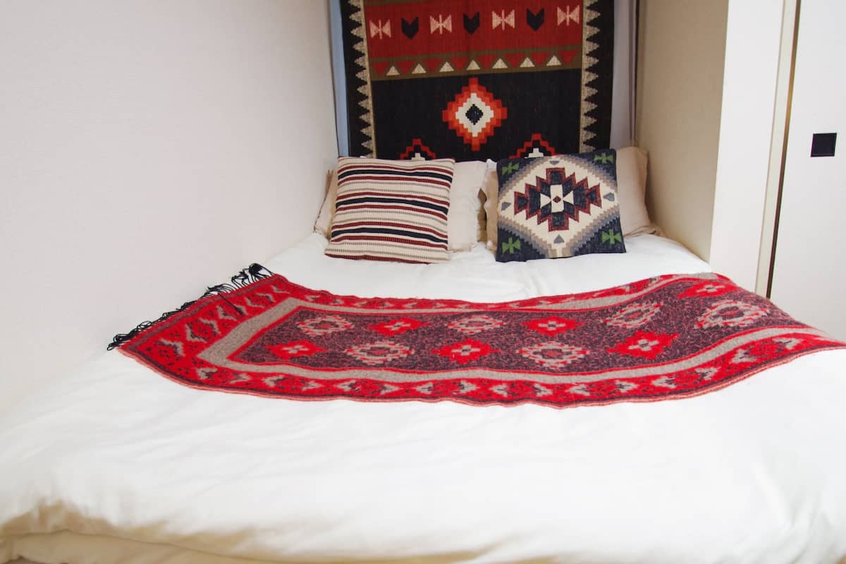 A bed with a white sheet, a red and black patterned blanket, and two pillows with geometric designs.