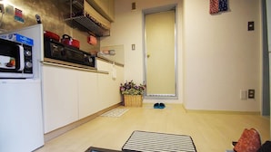 A compact kitchen with a microwave, stove, and a small refrigerator. There is a wooden floor, a door, and a basket with flowers. - suzuki4 (Tokyo)