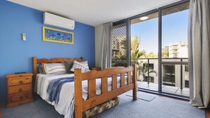 Un dormitorio con cama de madera, una mesita de noche, pared azul, unidad de aire acondicionado y vista al exterior por una puerta de cristal.