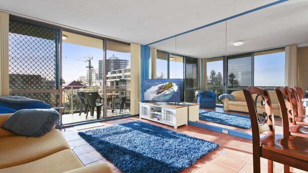 Un moderno salón con un televisor grande, alfombras azules y un comedor con sillas de madera.