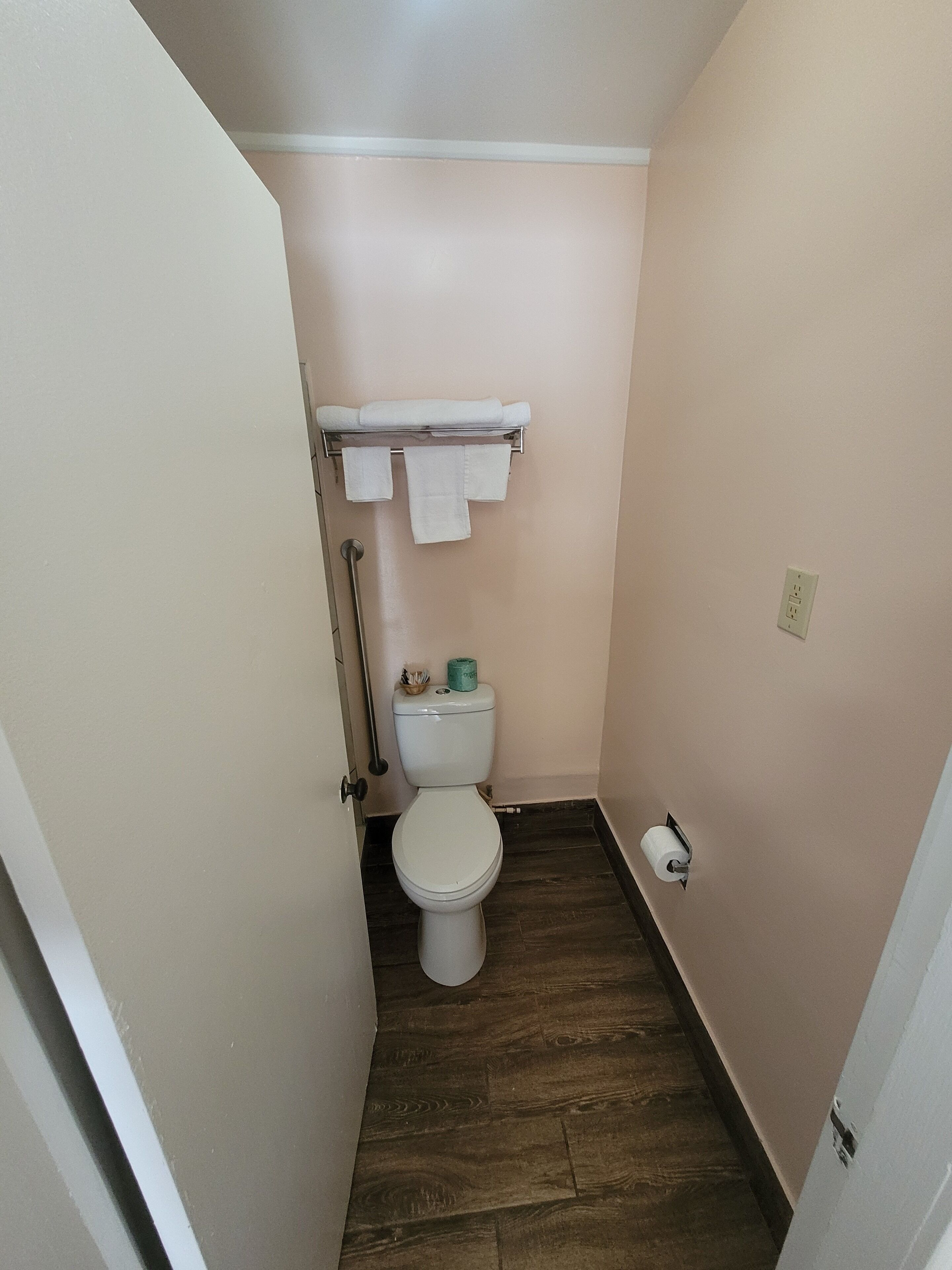 Un baño pequeño con inodoro, un perchero con toallas, dispensador de papel higiénico y piso de madera.