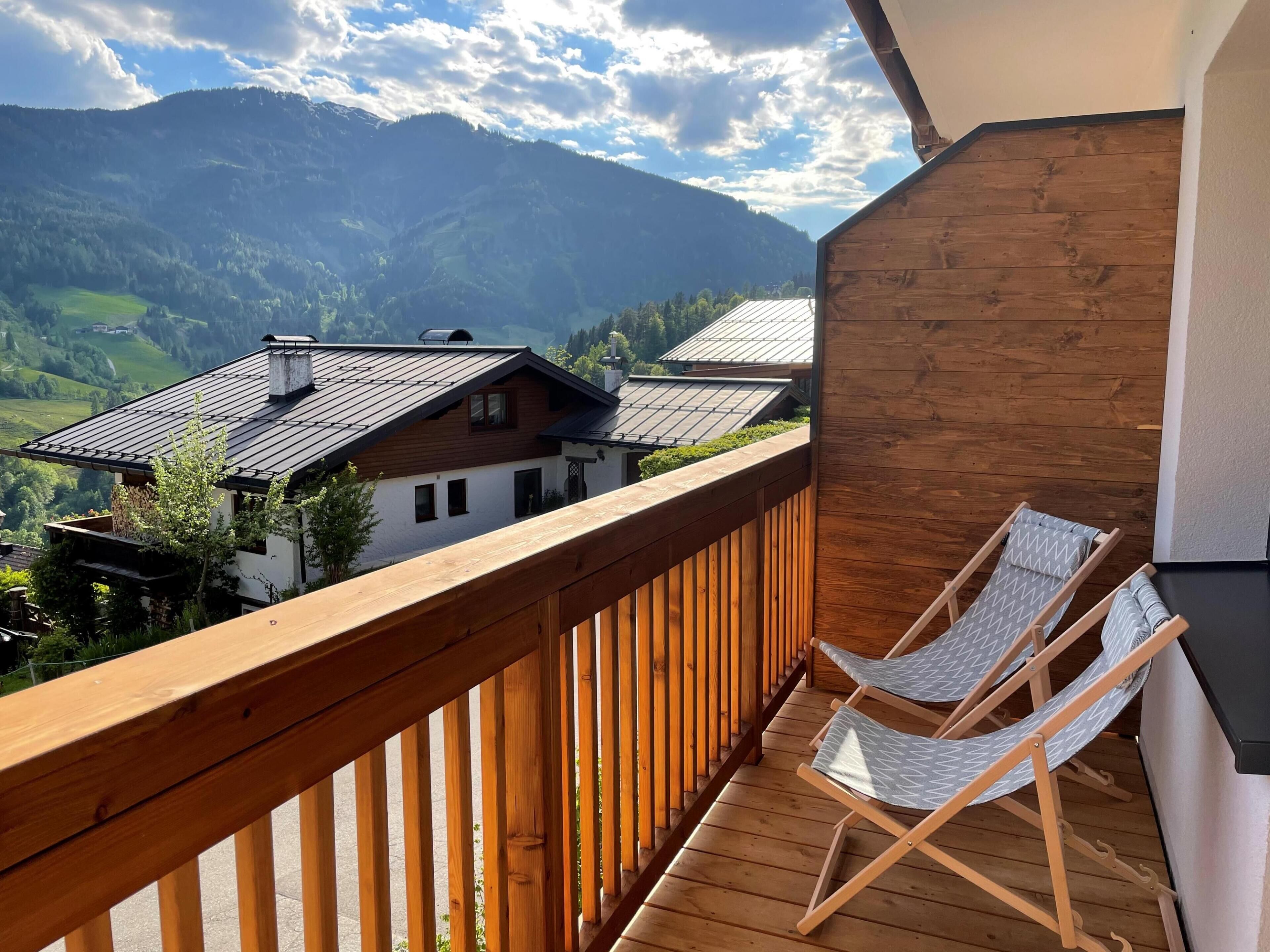 Een houten balkon met twee opklapbare stoelen, uitzicht op een berglandschap en een huis met schuin dak.