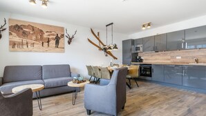 Una sala moderna con un sofá gris, una mesa de centro redonda y una zona de comedor con mesa y sillas de madera. Se ve un gran ciervo montado en la pared y esquíes. La cocina tiene armarios oscuros y una encimera de madera.