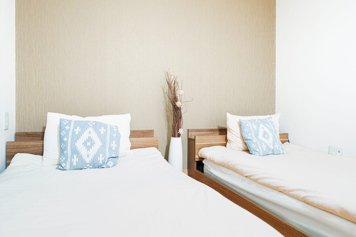 Dos camas individuales con almohadones blancos y azules estampados, un cabecero de madera y un jarrón blanco alto con plantas secas en el centro.