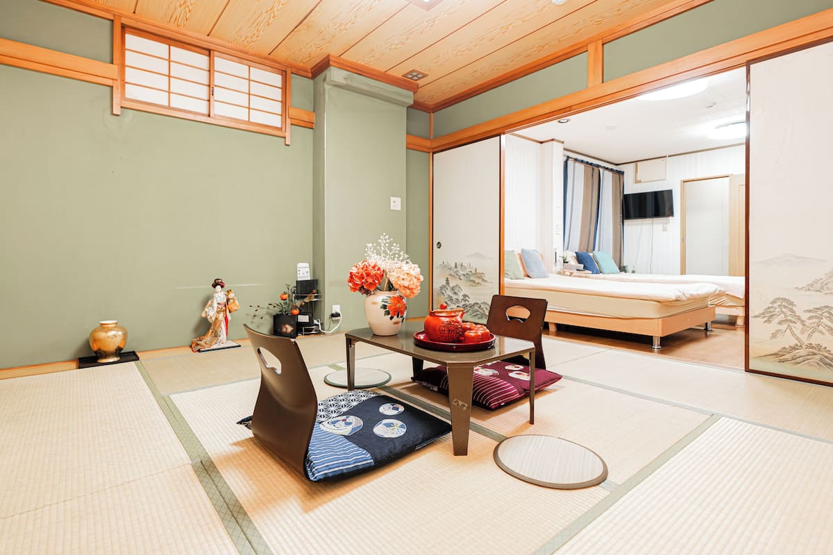 Una habitación japonesa tradicional con suelo de tatami, una mesa baja con un florero y una cama con futón, además de puertas correderas.