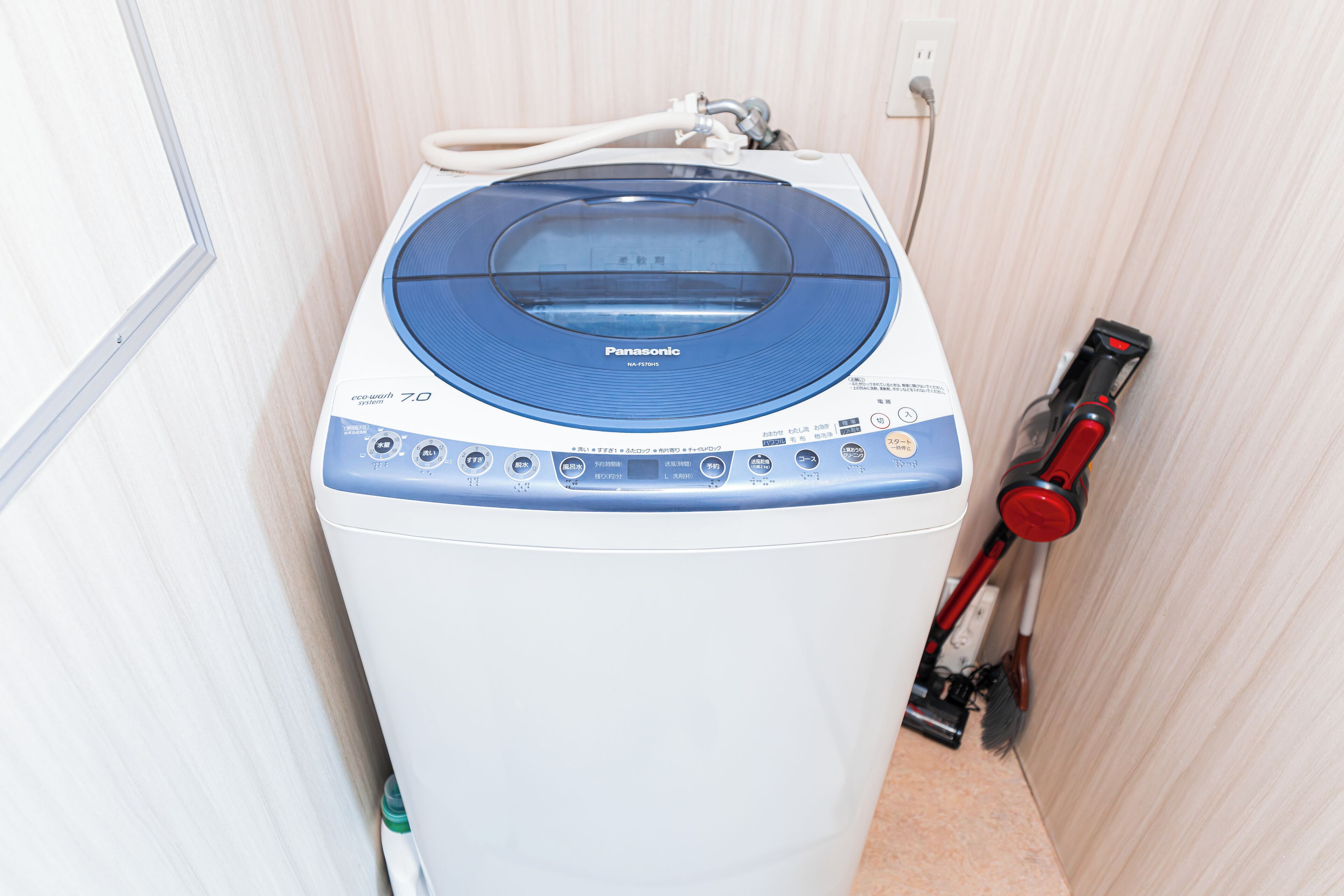 Une machine à laver compacte, blanche et bleue, dotée d’un couvercle transparent, située dans une petite pièce aux murs en bois et où une prise de courant est visible.