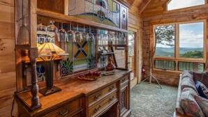 Accogliente interno di una baita con arredamento in legno, una finestra a vetri colorati, un comò in legno con bicchieri e vista sull'esterno.