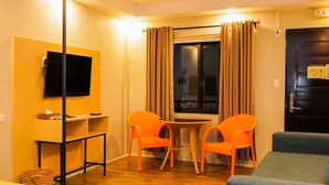Une chambre d’hôtel compacte avec un plancher en bois, un bureau doté d’un téléviseur à écran plat, deux fauteuils orange, un canapé bleu et une fenêtre avec des rideaux.