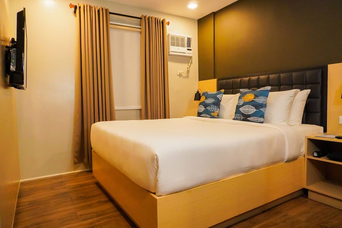 A hotel room with a bed, a bedside table, a TV, an air conditioner, and curtains.