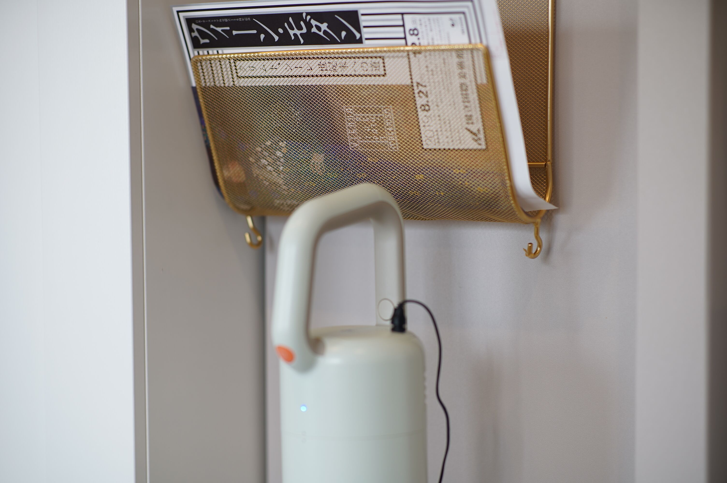 A white vacuum cleaner with a handle and cord, mounted on a wall with a metal mesh shelf above it.