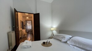 A hotel room with a bed, a bedside lamp, a radiator, and a hallway leading to another room.