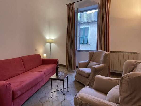 A cozy living room with a red sofa, beige armchairs, a small table with a bottle and glasses, a floor lamp, and a window with curtains.