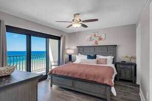 A bedroom with a bed, a ceiling fan, a nightstand, a lamp, and a view of the ocean.