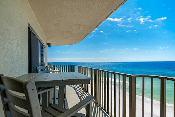 A balcony with a table and chairs, overlooking a beach with clear blue water and a bright sky.