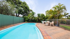 A swimming pool with a brick border, surrounded by a green fence, and a patio area with outdoor furniture. - Croydon Ave 50 - Currimundi (CURRIMUNDI)