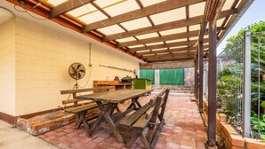 An outdoor patio with wooden benches and a fan, a brick floor, and a green fence enclosing the area. - Croydon Ave 50 - Currimundi (CURRIMUNDI)