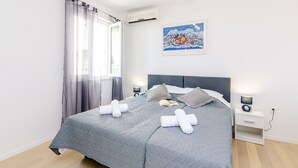 A neatly made bed with a grey quilt, two rolled towels, and pillows on a white nightstand with a lamp. - Miracle Apartments (Dubrovnik)