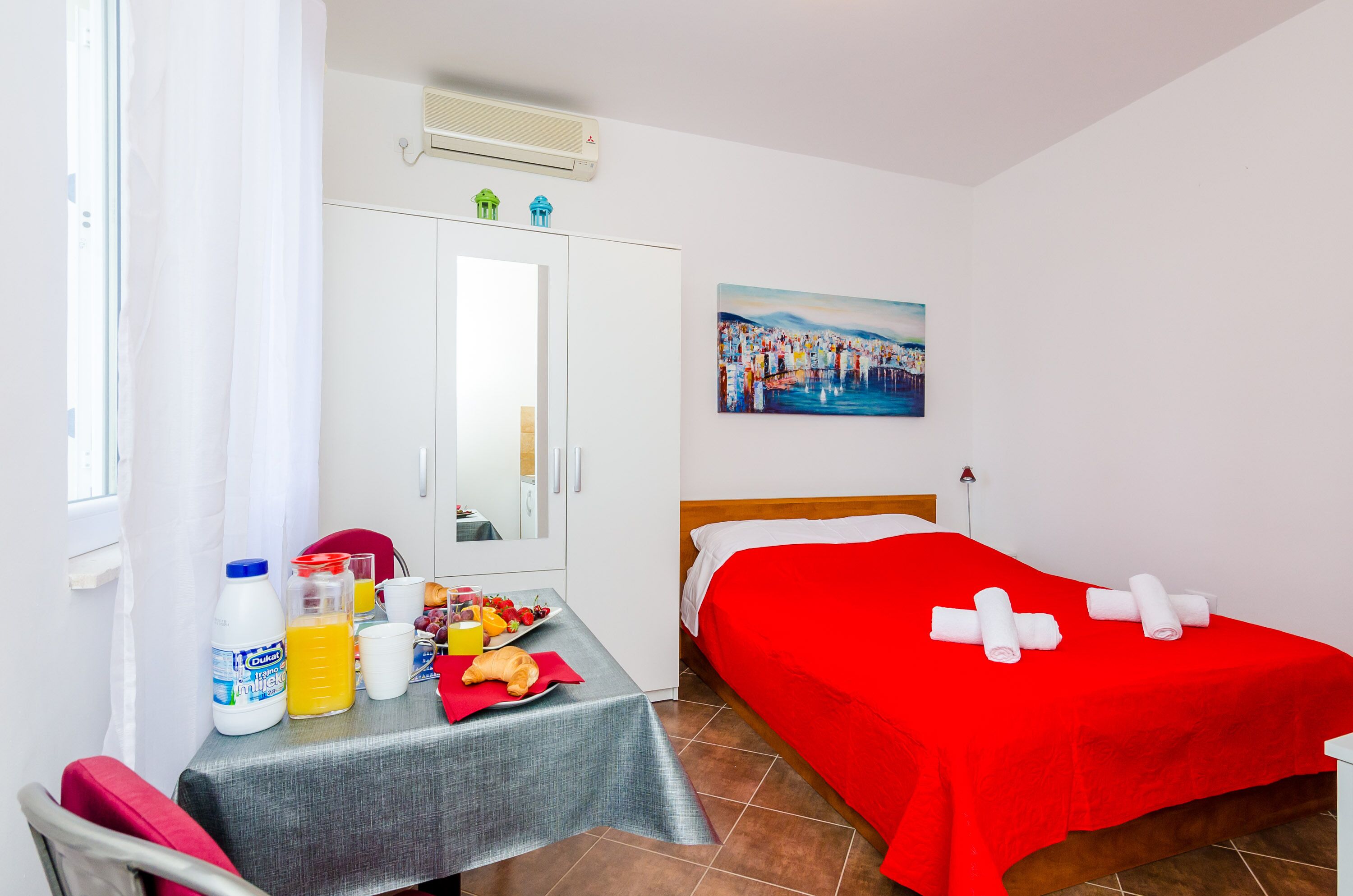 Una habitación de hotel con una cama, una mesa con alimentos para el desayuno, un cuadro en la pared y una ventana con cortinas.